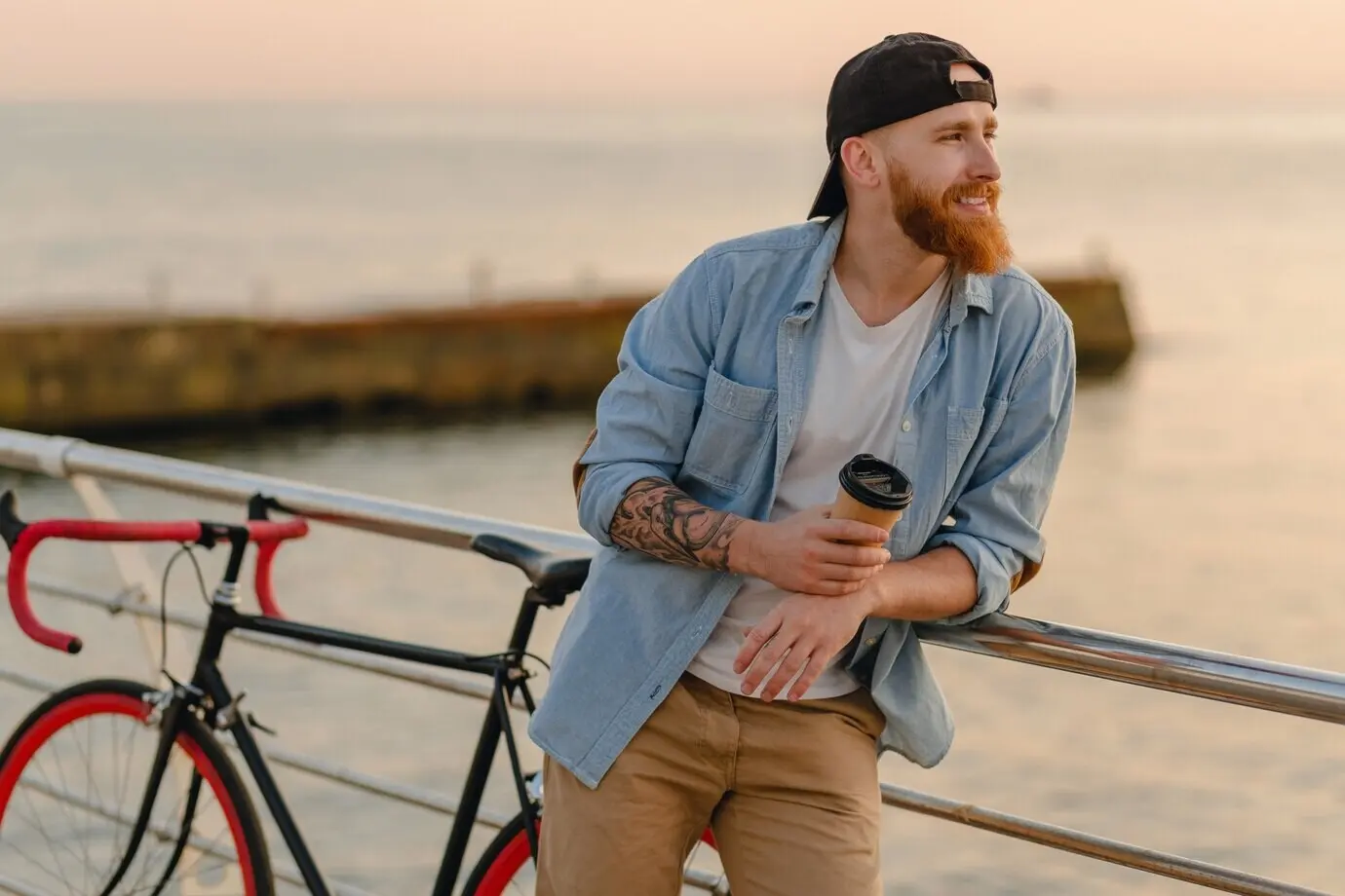 Gutaussehender, lächelnder Mann im Hipster-Stil mit rotem Bart, in Jeanshemd und Kappe, mit Fahrrad, der beim morgendlichen Sonnenaufgang am Meer Kaffee trinkt, Reisender mit gesundem, aktivem Lebensstil.