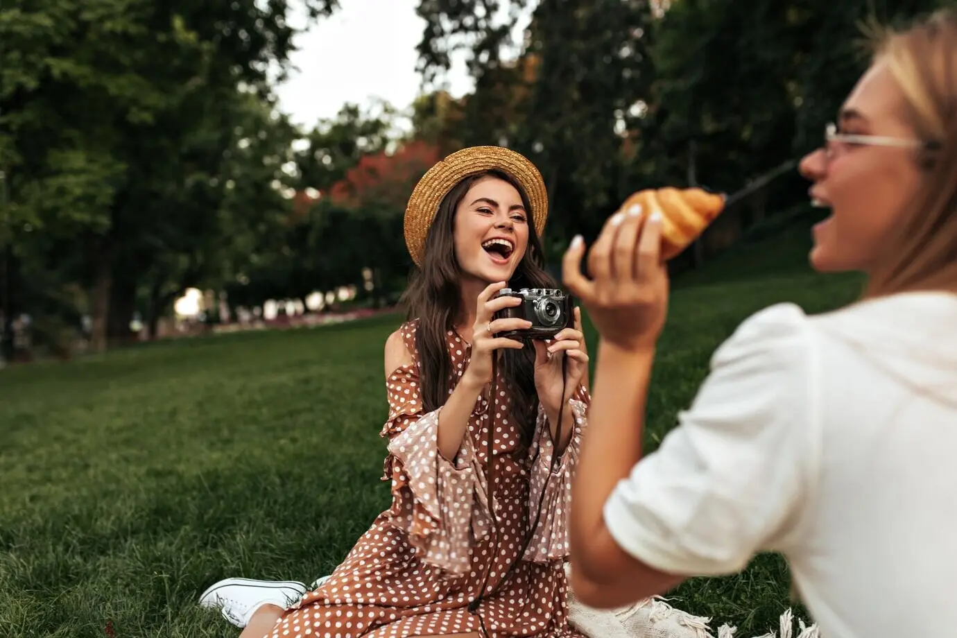 Coole junge brünette Frau mit Boater-Hut und gepunktetem Kleid lacht und fotografiert ihre Freundin mit einer Retro-Kamera. Blondes Mädchen im weißen Outfit hält einen Apfel und macht ein Picknick. Unschärfe-Effekt.