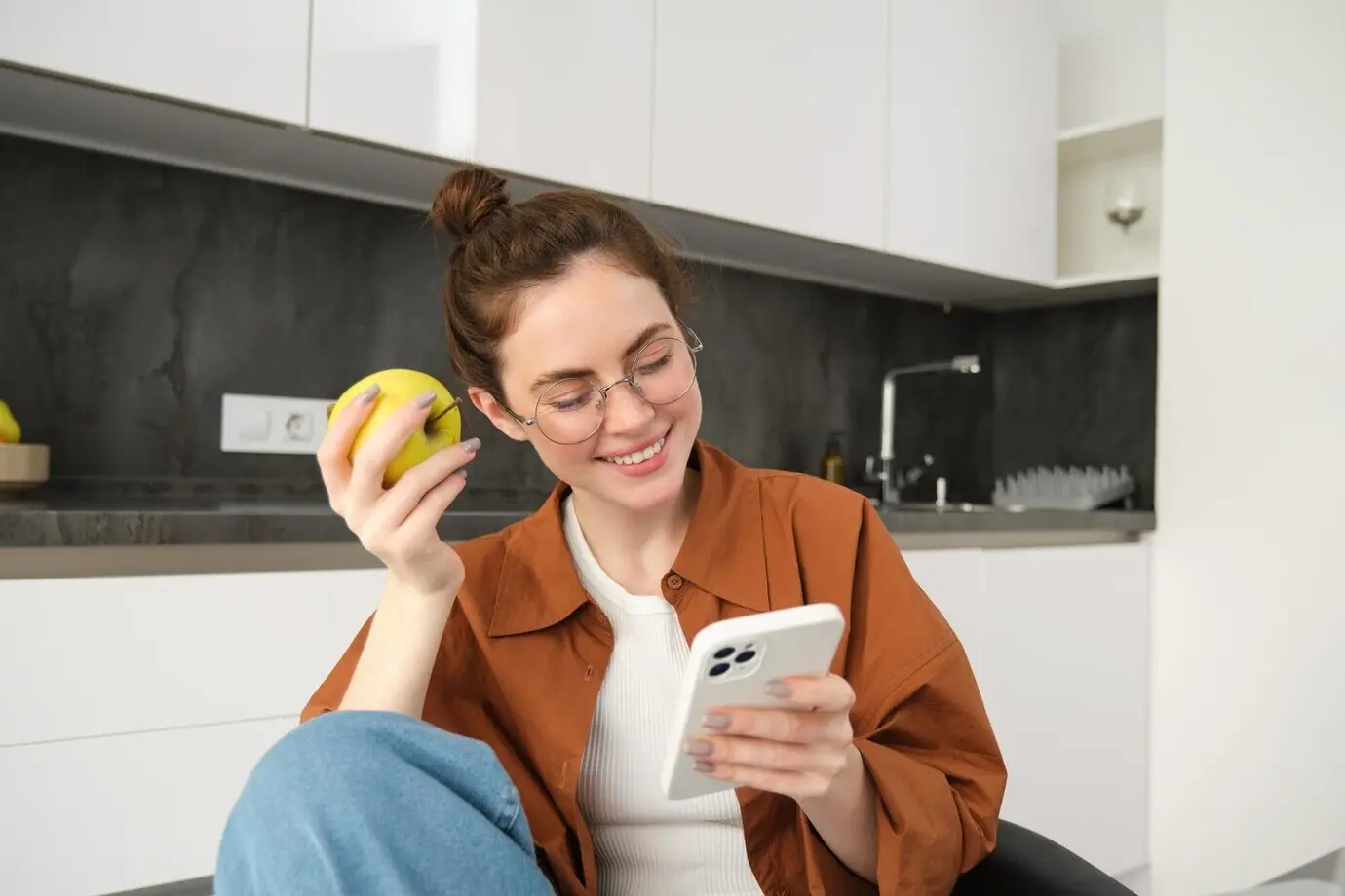 Eine junge Frau entspannt zu Hause, blickt auf ihr Smartphone, liest Nachrichten in sozialen Medien und isst etwas Grünes.